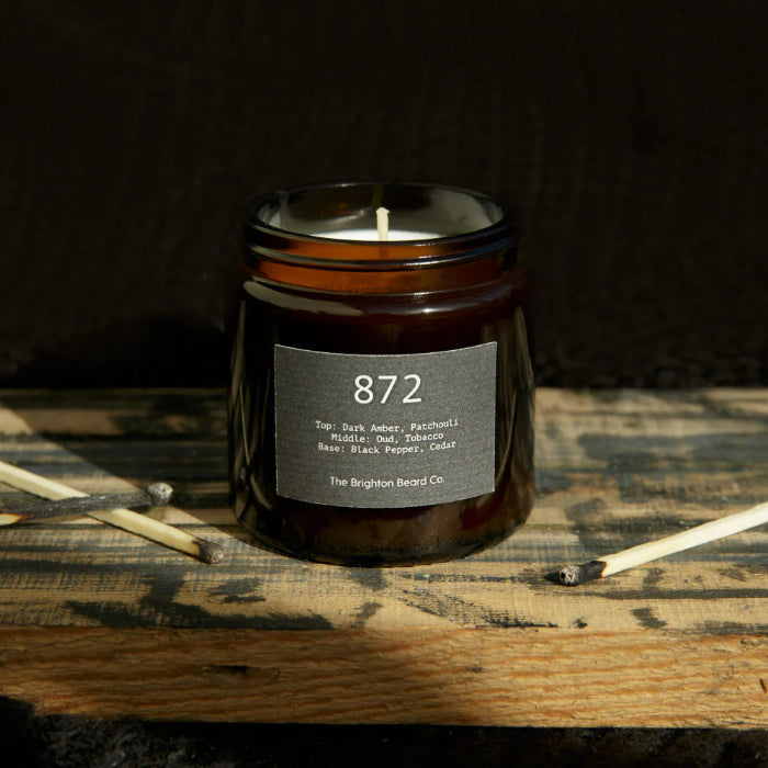 Candle in a brown jar with a label on a wooden surface, featuring the Brighton Beard Co brand.