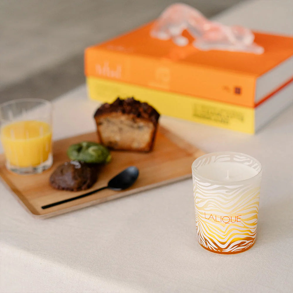 Candle with 'Lalique' branding on a table with a book, glass of orange juice, and pastries.