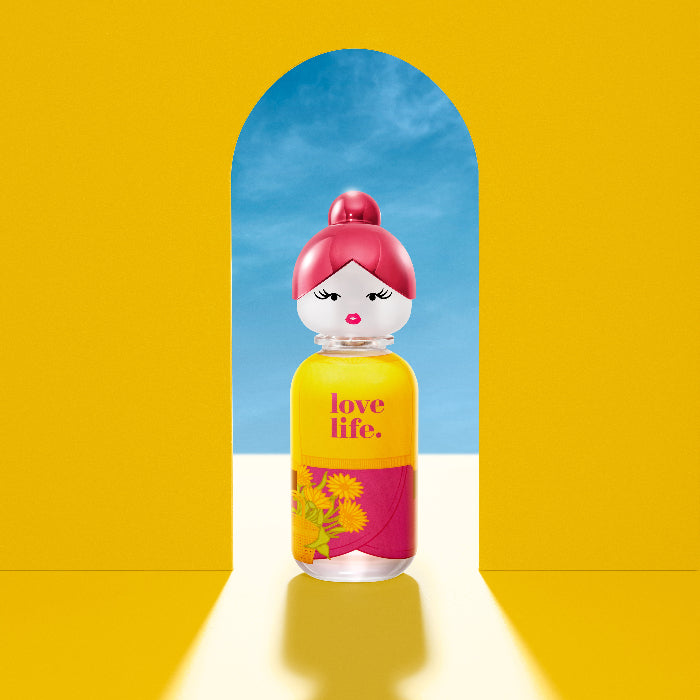 Perfume bottle with pink cap and yellow label featuring 'love life.' text, set against a bright yellow background with a blue archway.