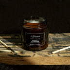 Candle in a jar labeled 'Alfriston' on a wooden surface with a dark background