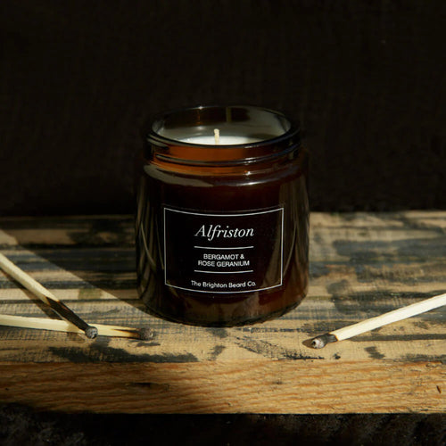 Candle in a jar labeled 'Alfriston' on a wooden surface with a dark background